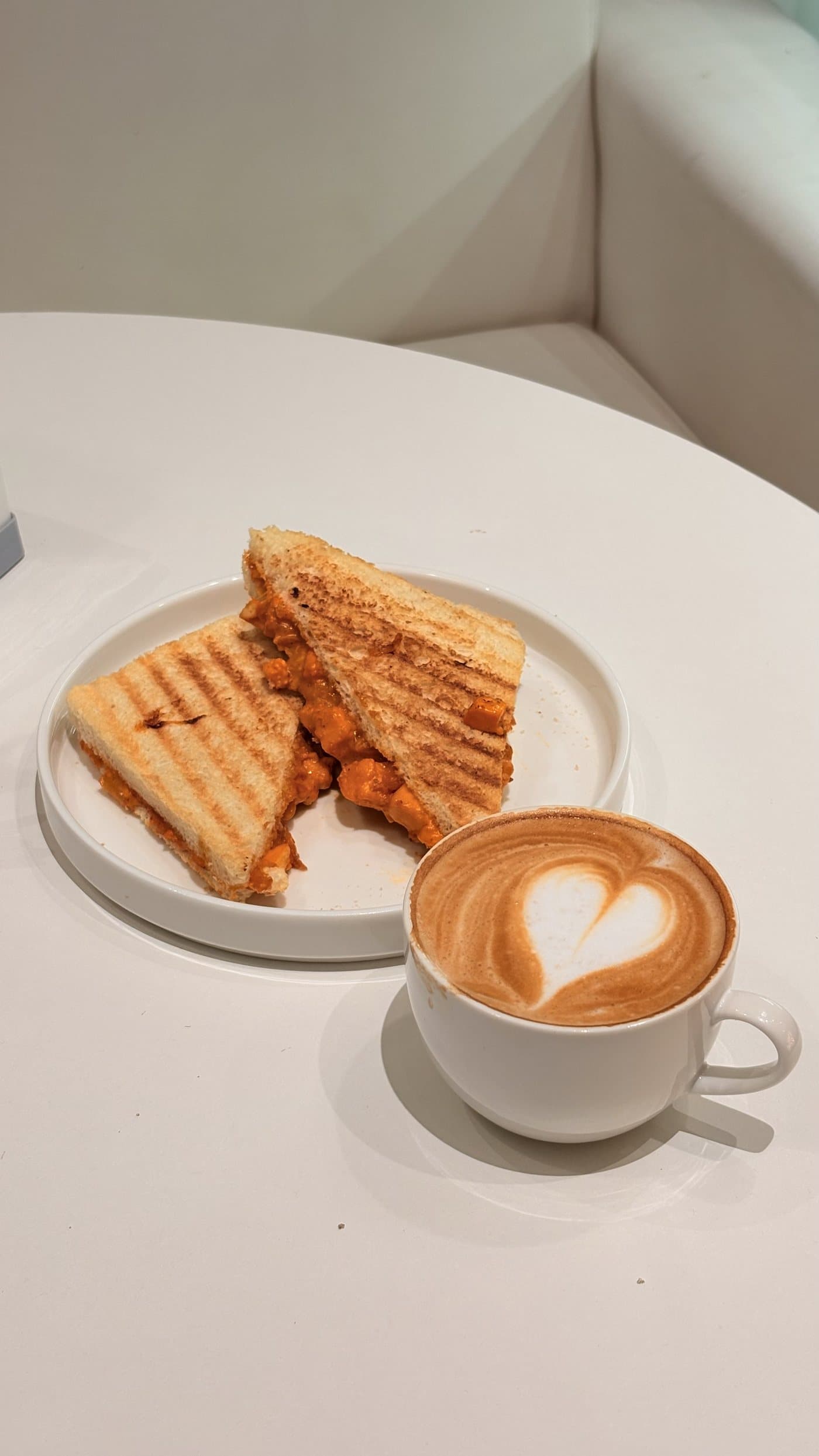 A cappuccino with heart latte art beside a toasted sandwich on a white café table.